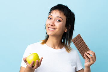 Canvas Print - Young Uruguayan woman over isolated blue background taking a chocolate tablet in one hand and an apple in the other