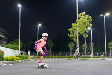 asian child or kid girl playing surf skate or skateboard in skating rink or sport public park at night to wearing face mask and helmet with elbow pads wrist and knee support for body safety to protect