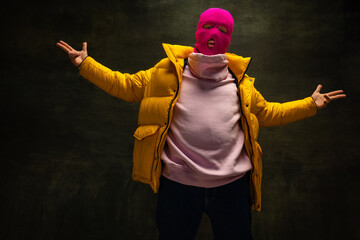 Studio shot of young anonymous man wearing pink balaclava and yellow down jacket, coat isolated on dark vintage background. Concept of safety, art, fashion