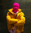 Studio shot of young anonymous man wearing pink balaclava and yellow down jacket, coat isolated on dark vintage background. Concept of safety, art, fashion