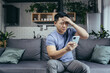 © Liubomir - Man reading bad news from phone, asian frustrated and sad looking at phone screen sitting on sofa at home
