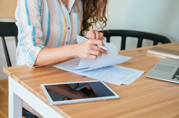 Close up photo of woman hands signing a contract papers or writing something on a document while sitting at home office desk with laptop computer and digital tablet