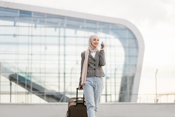 Poster - Air flight journey and business concept. Pretty traveler tourist arabian woman in hijab carrying suitcase and talking phone, walking near modern airport terminal