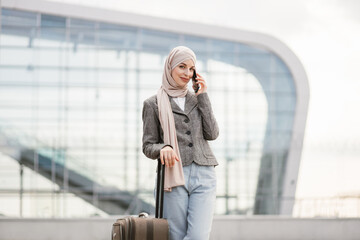 Wall Mural - Beautiful young excited woman in hijab talking on the phone with friend, in anticipation of a wonderful trip and vacation. Arab lady standing with suitcase near airport