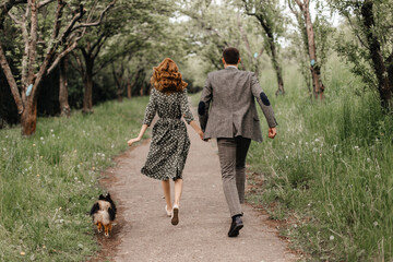 a couple in retro style running along a garden path with a small dog behind them