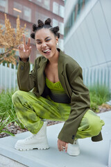 Wall Mural - Joyful millennial girl squats winks eye and sticks out tongue shows peace gesture dressed in dark green jacket trousers and white boots feels self confident poses in urban place. Hey look at me