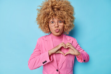 Wall Mural - Romantic curly haired young woman dressed in formal pink jacket makes heart gesture keeps lips folded loves someone isolated over blue background. I love you. St Valentines Day. Body language