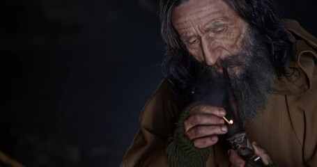 Wall Mural - fantastic monk sorcerer with a gray beard chids in a cave and lights a pipe with tobacco. close up.