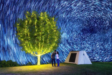 Wall Mural - couple under a tree adorn lights beside tent camping at night. there are beautiful stars trail in th