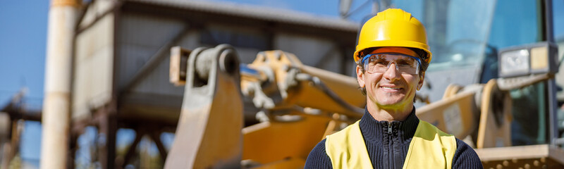 Joyful matured man engineer wearing safety helmet, glasses and vest while keeping arms crossed and smiling