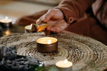 Wall Mural - Woman hands burning Palo Santo, before ritual on the table with candles and green plants. Smoke of smudging treats pain and stress, clears negative energy and meditation wooden stick