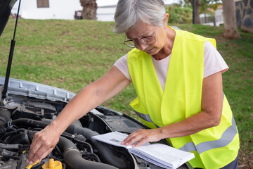 Senior driver stopped in the street for a car breakdown with the hood open wearing the yellow vest waiting for roadside assistance. Look at the instruction booklet to understand what is broken