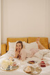 © Look! - Cheerful young caucasian girl licks cream with fingers while lying on bed in cozy bright room. Blonde in silk pajamas spends morning in good insistence. Weekend concept