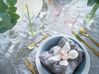 Colorful tableware and relaxed table setting with flowers. Grey linen table cloth and a napkin, golden cutlery and colorful tableware. Orchid flower on top of a napkin.