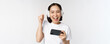 Happy asian girl gamer, playing on mobile phone, watching on smartphone, wearing headphones, standing over white background