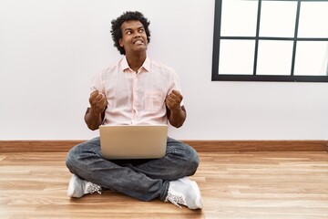 Poster - African man with curly hair using laptop sitting on the floor very happy and excited doing winner gesture with arms raised, smiling and screaming for success. celebration concept.