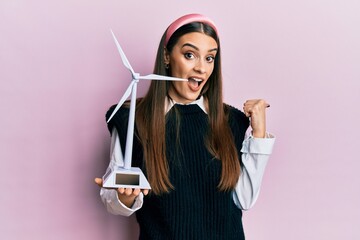 Wall Mural - Beautiful brunette young woman holding solar windmill for renewable electricity pointing thumb up to the side smiling happy with open mouth