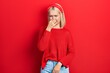 Beautiful blonde woman wearing casual red sweater smelling something stinky and disgusting, intolerable smell, holding breath with fingers on nose. bad smell