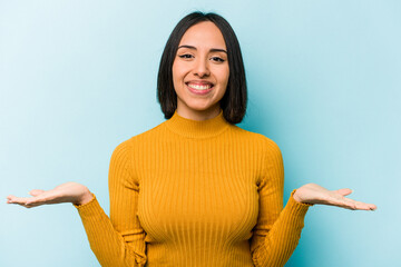 Young hispanic woman isolated on blue background makes scale with arms, feels happy and confident.