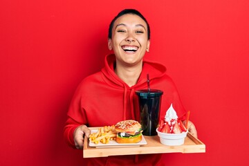 Sticker - Beautiful hispanic woman with short hair eating a tasty classic burger with fries and soda smiling and laughing hard out loud because funny crazy joke.