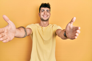 Wall Mural - Young hispanic man wearing casual t shirt looking at the camera smiling with open arms for hug. cheerful expression embracing happiness.