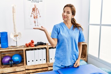 Sticker - Middle age hispanic physiotherapist woman working at pain recovery clinic smiling cheerful presenting and pointing with palm of hand looking at the camera.