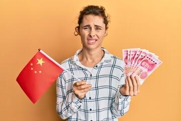 Poster - Young brunette woman holding china flag and yuan banknotes clueless and confused expression. doubt concept.