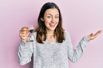 Young brunette woman holding sand clock celebrating achievement with happy smile and winner expression with raised hand