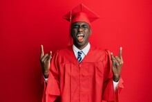 Young African American Man Wearing Graduation Cap And Ceremony Robe Shouting With Crazy Expression Doing Rock Symbol With Hands Up. Music Star. Heavy Concept.