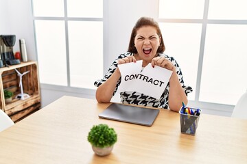 Poster - Middle age hispanic woman breaking contract paper at the office angry and mad screaming frustrated and furious, shouting with anger. rage and aggressive concept.