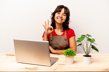 Wall Mural - Young gardener hispanic woman sitting on a table isolated on white background cheerful and confident showing ok gesture.