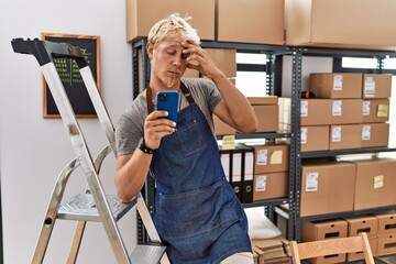 Wall Mural - Young blond man using smartphone working at storehouse worried and stressed about a problem with hand on forehead, nervous and anxious for crisis