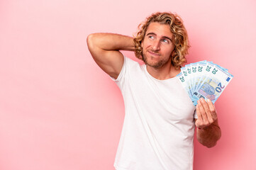 Wall Mural - Young caucasian man holding banknotes isolated on pink background touching back of head, thinking and making a choice.