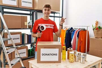 Wall Mural - Young caucasian volunteer man packing donations box at charity center smiling happy pointing with hand and finger to the side