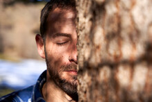 Man With Eye Closed Standing Behind Tree Trunk On Sunny Day