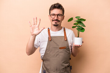 Wall Mural - Young caucasian gardener man holding a plant isolated on beige background smiling cheerful showing number five with fingers.