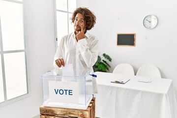 Sticker - Young hispanic man voting putting envelop in ballot box pointing to the eye watching you gesture, suspicious expression