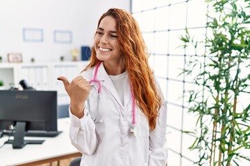 Poster - Young redhead woman wearing doctor uniform and stethoscope at the clinic smiling with happy face looking and pointing to the side with thumb up.