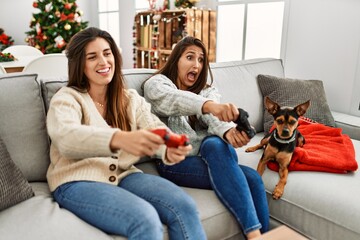 Two women playing video game sitting with dog by christmas tree at home