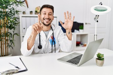 Wall Mural - Young doctor working at the clinic using computer laptop showing and pointing up with fingers number six while smiling confident and happy.