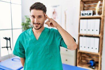 Wall Mural - Young physiotherapist man working at pain recovery clinic smiling pointing to head with one finger, great idea or thought, good memory