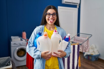 Poster - Young hispanic woman holding detergent bottles sticking tongue out happy with funny expression.