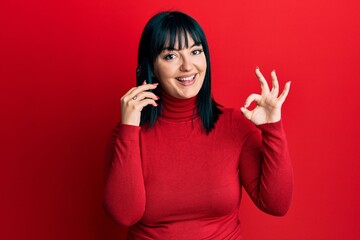 Sticker - Young hispanic woman having conversation talking on the smartphone doing ok sign with fingers, smiling friendly gesturing excellent symbol