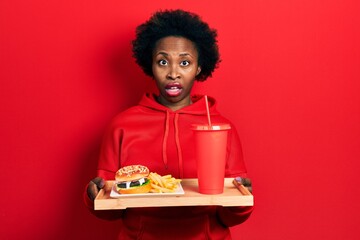 Poster - Young african american woman eating a tasty classic burger with fries and soda in shock face, looking skeptical and sarcastic, surprised with open mouth