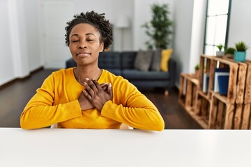 Sticker - Young african american woman wearing casual clothes sitting on the table at home smiling with hands on chest with closed eyes and grateful gesture on face. health concept.