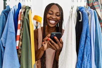 Wall Mural - Young african american customer woman smiling happy appearing through clothes using smartphone at clothing store.