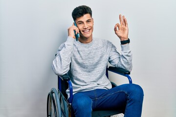 Canvas Print - Young hispanic man sitting on wheelchair talking on the smartphone doing ok sign with fingers, smiling friendly gesturing excellent symbol