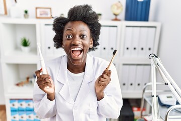 Wall Mural - African dentist woman holding electric toothbrush and normal toothbrush celebrating crazy and amazed for success with open eyes screaming excited.