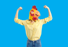 Woman In Funny Chicken Disguise Pretends To Be Super Strong. Portrait Of Confident Young Girl Wearing Strange Weird Ridiculous Absurd Bizarre Hallowen Mask Flexing Her Arms Isolated On Blue Background