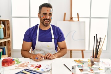 Poster - Young hispanic man with beard at art studio with painted face winking looking at the camera with sexy expression, cheerful and happy face.